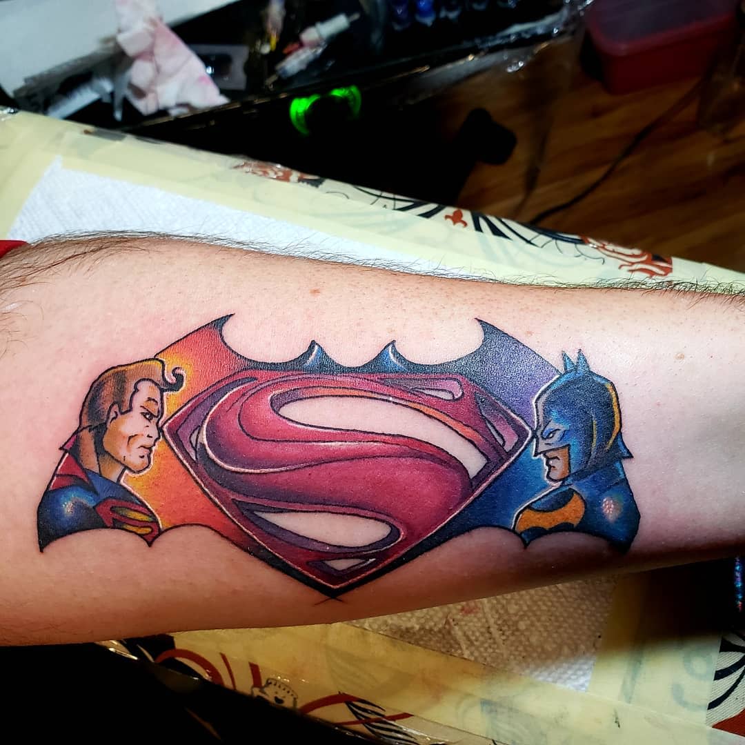 Tattoo featuring Superman and Batman faces with a combined "S" and bat symbol on an arm